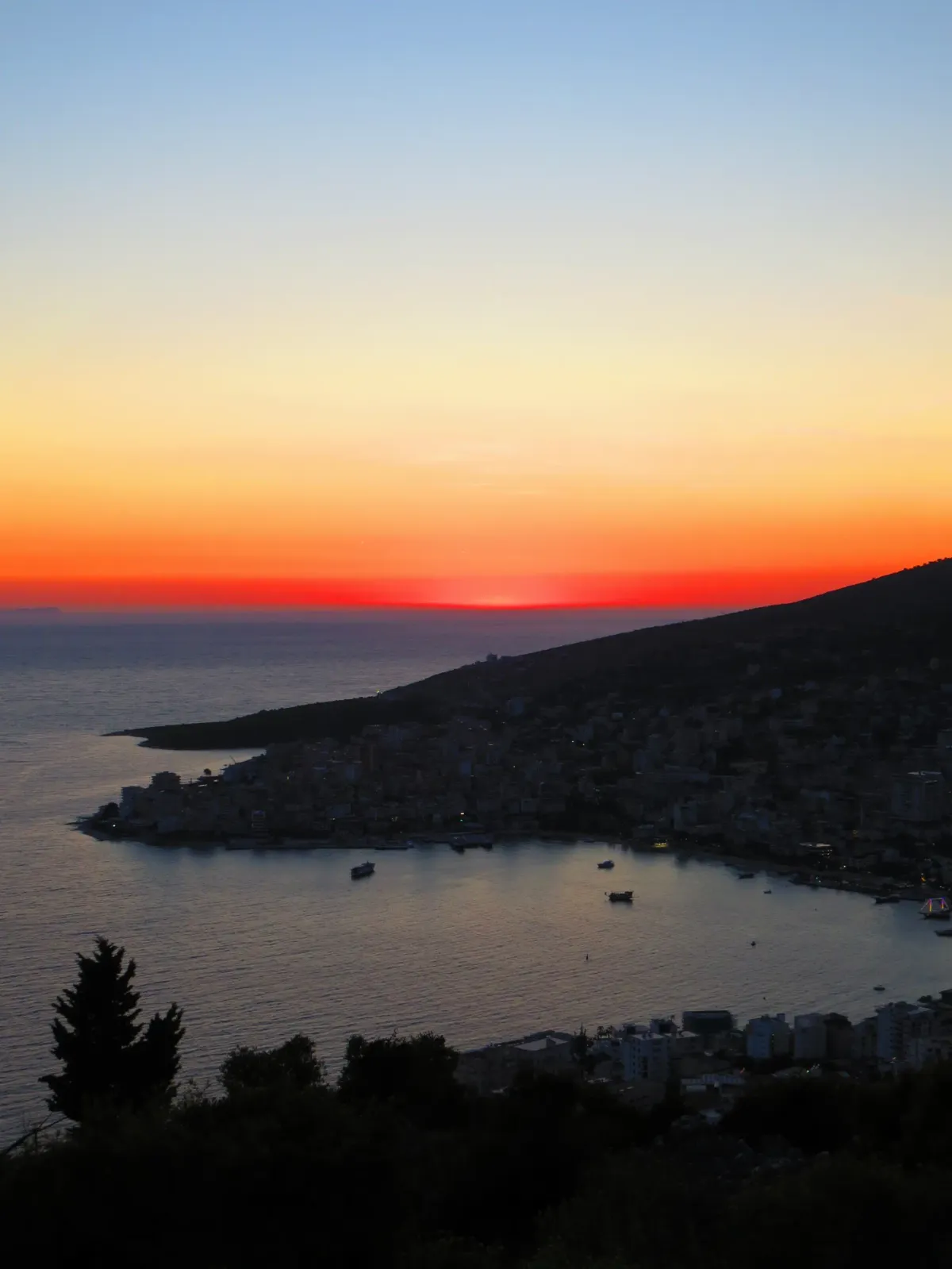 The sun sets over the bay of Sarandë, its glow spreading across the Ionian Sea and the hillside town below.