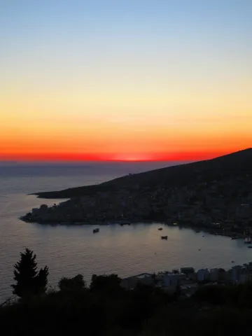 The sun sets over the bay of Sarandë, its glow spreading across the Ionian Sea and the hillside town below.