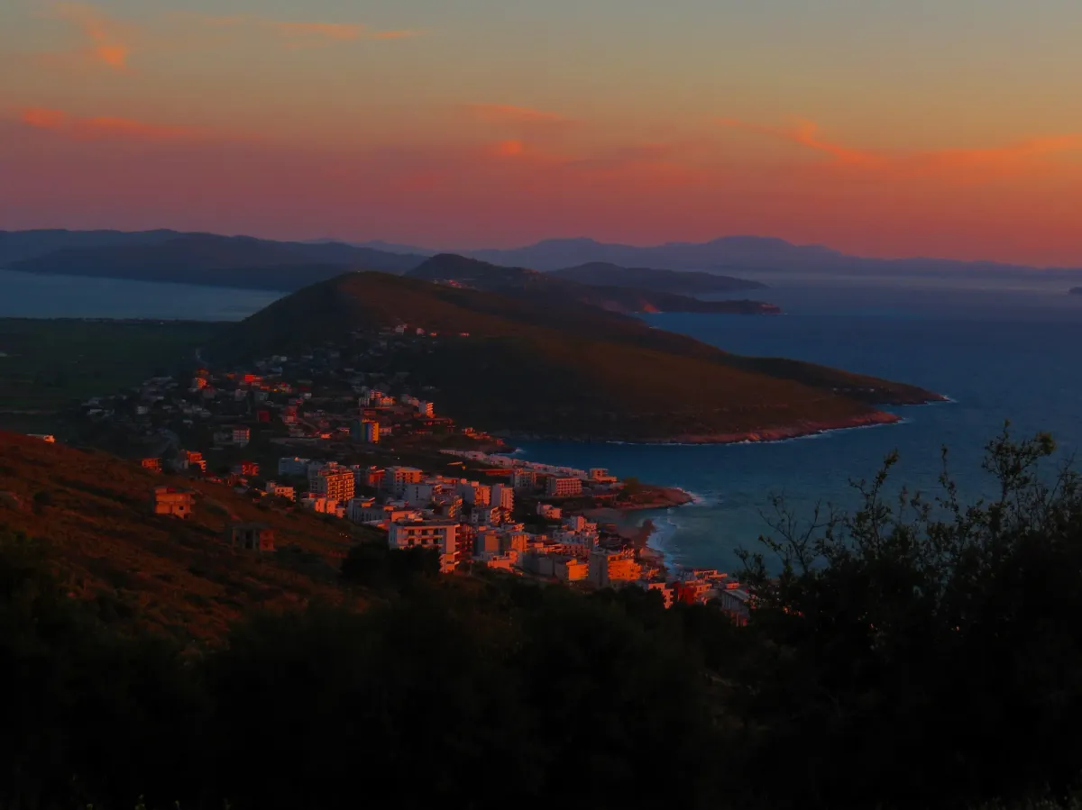 A panoramic view of Sarandë's coastline and hills glowing under a pastel sunset.