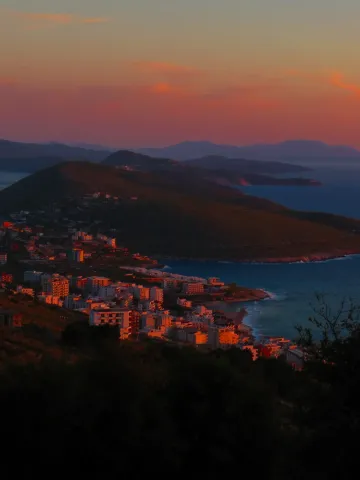 A panoramic view of Sarandë's coastline and hills glowing under a pastel sunset.