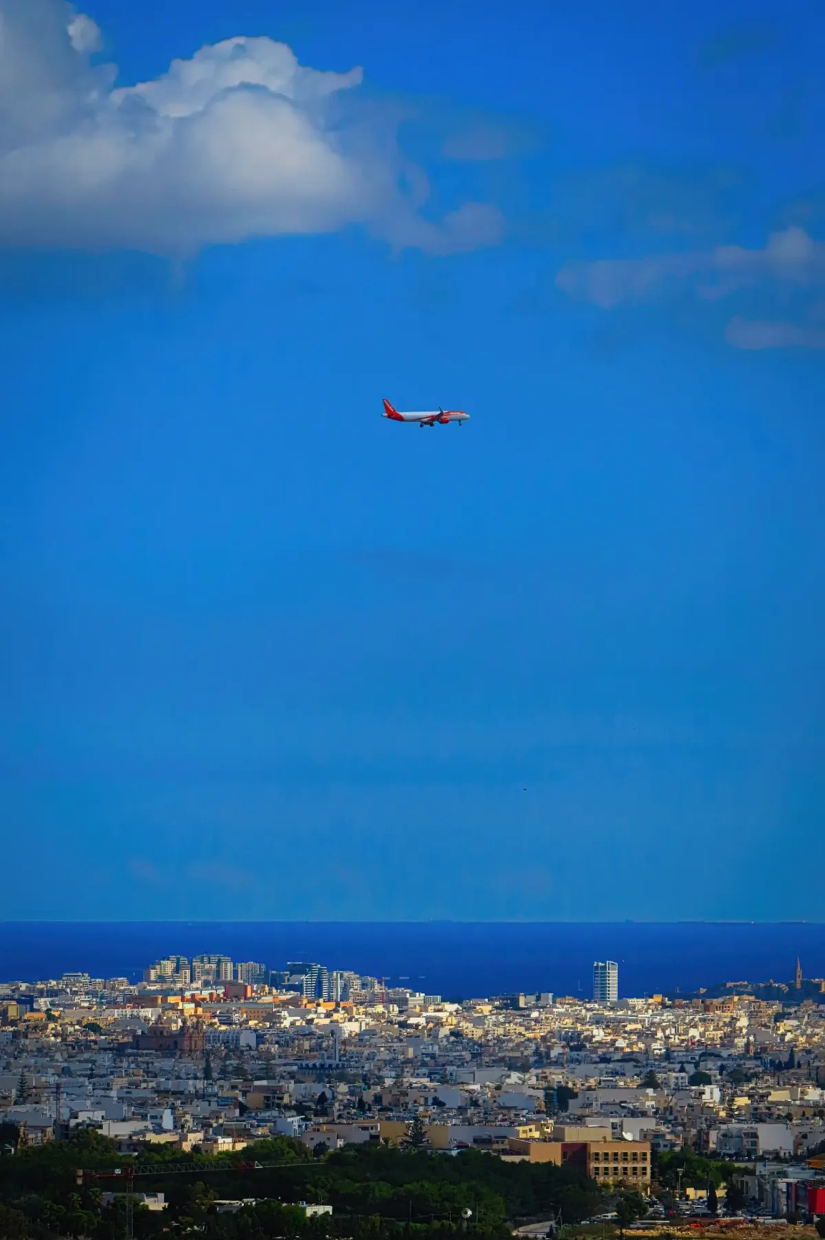 A passenger jet descends over the coastline of Malta under a deep blue sky.