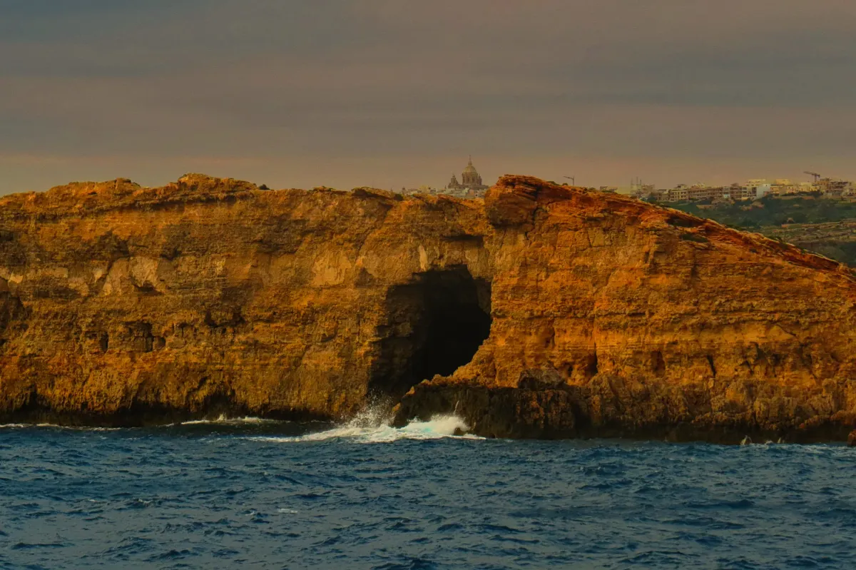 Waves crash into a sea cave beneath sunlit cliffs, with a church dome visible on the horizon.