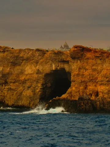 Waves crash into a sea cave beneath sunlit cliffs, with a church dome visible on the horizon.